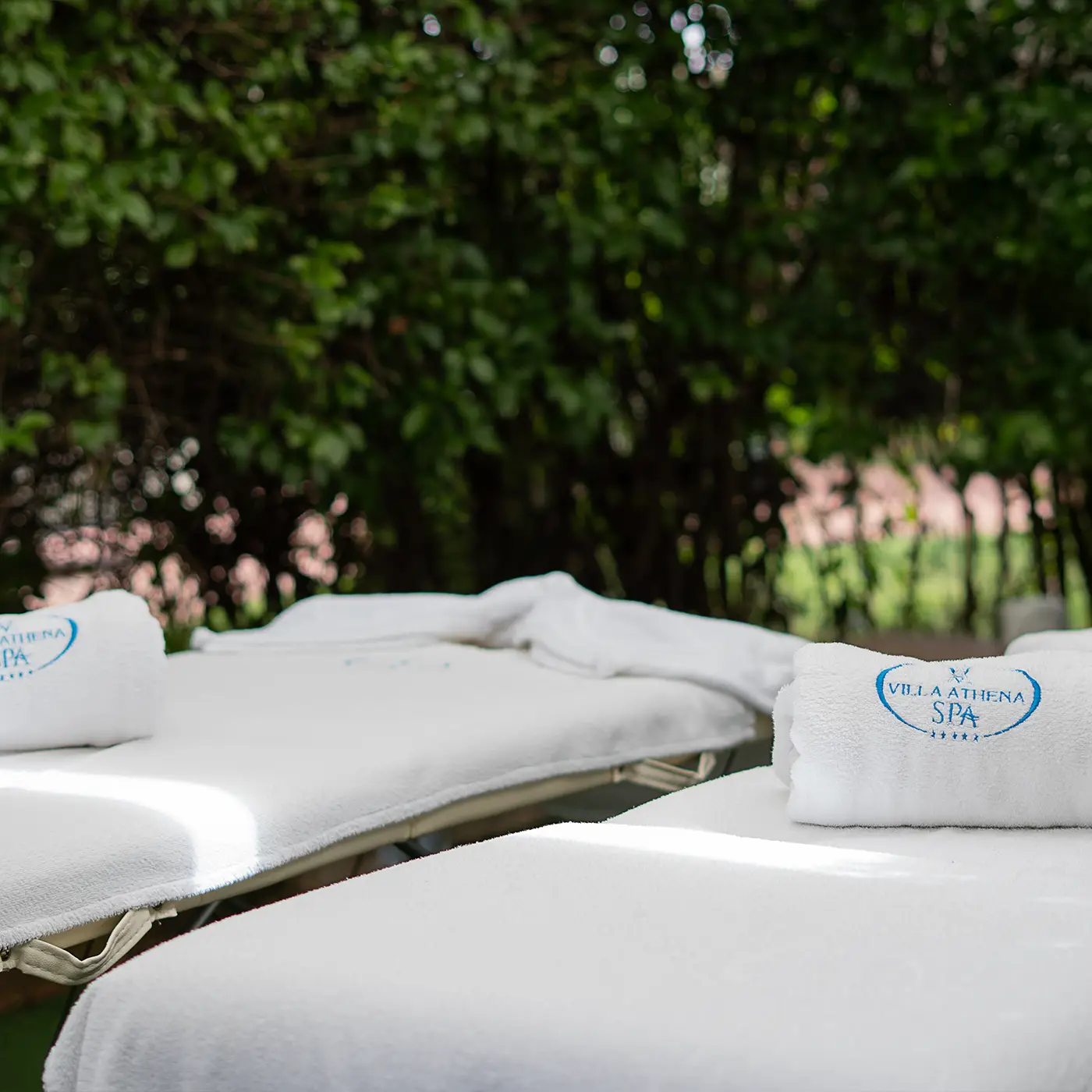 Lettini da massaggio all'aperto con asciugamani bianchi e logo Villa Athena SPA, immersi nel verde del giardino dell'hotel.