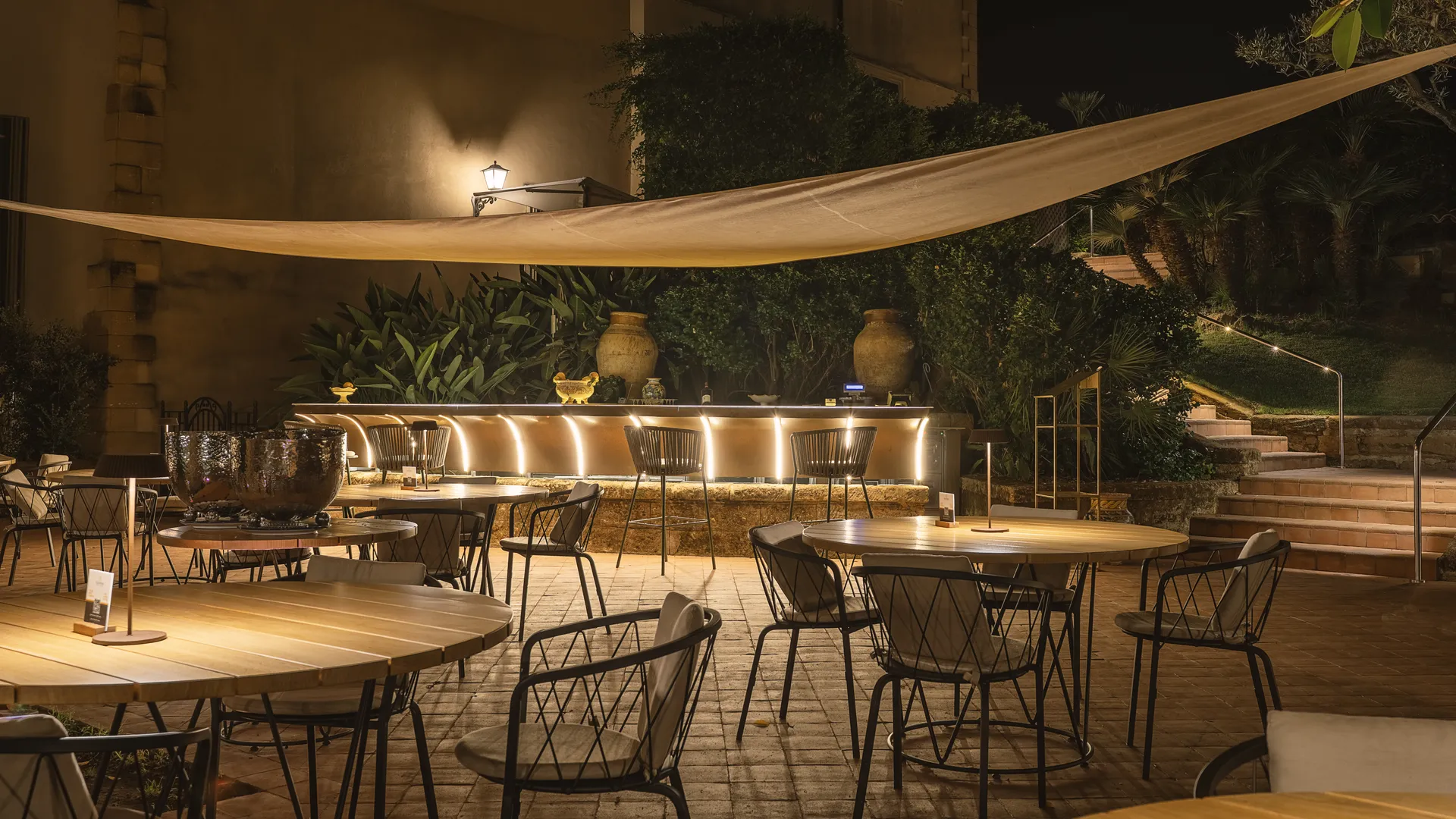 Atmosfera serale del Ristorante Federico II con tavoli illuminati, banco bar retroilluminato e vele parasole nel giardino.
