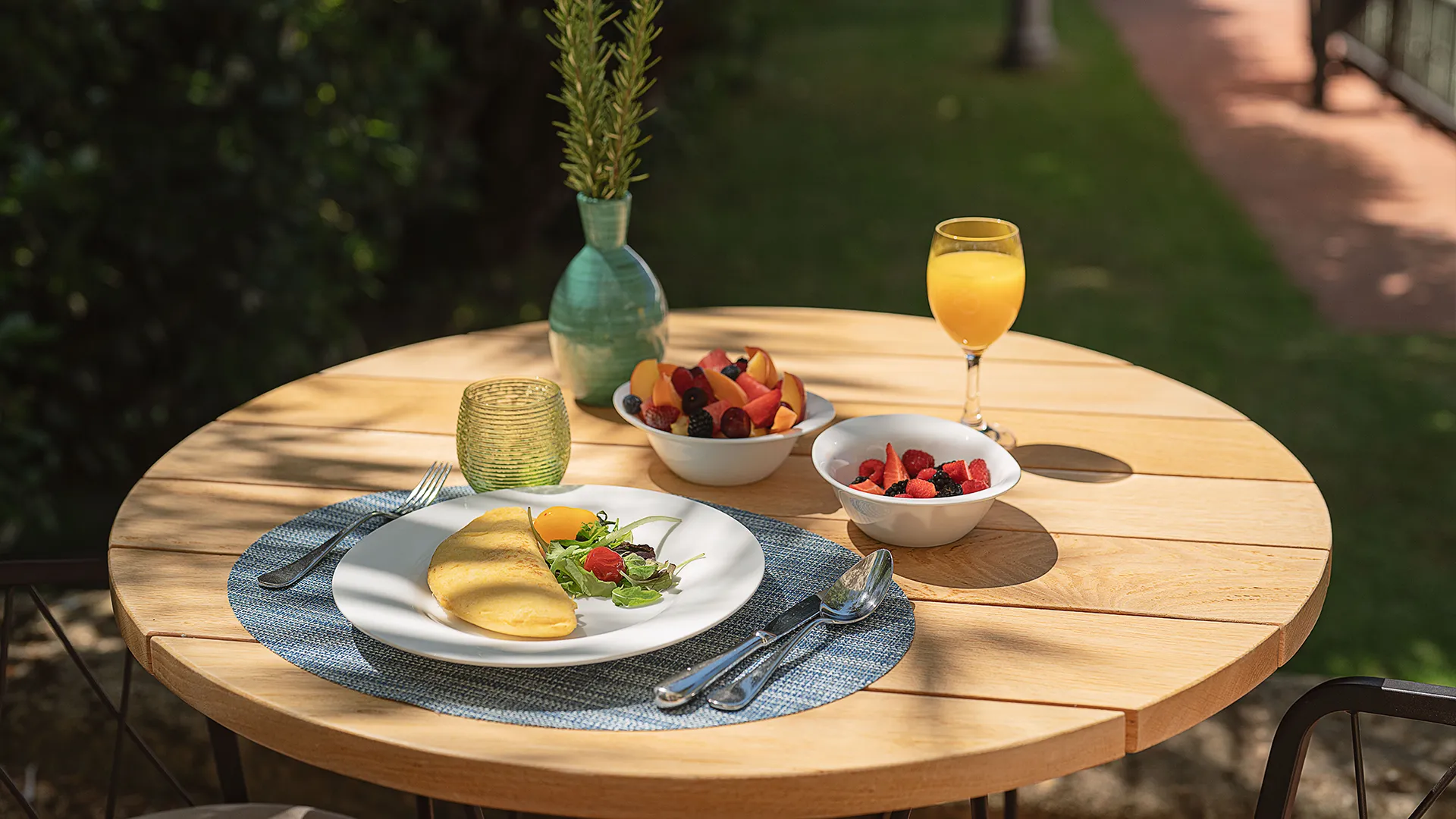 Colazione all'aperto servita su un tavolo in legno con omelette, insalata, macedonia fresca, frutti di bosco e succo d'arancia.
