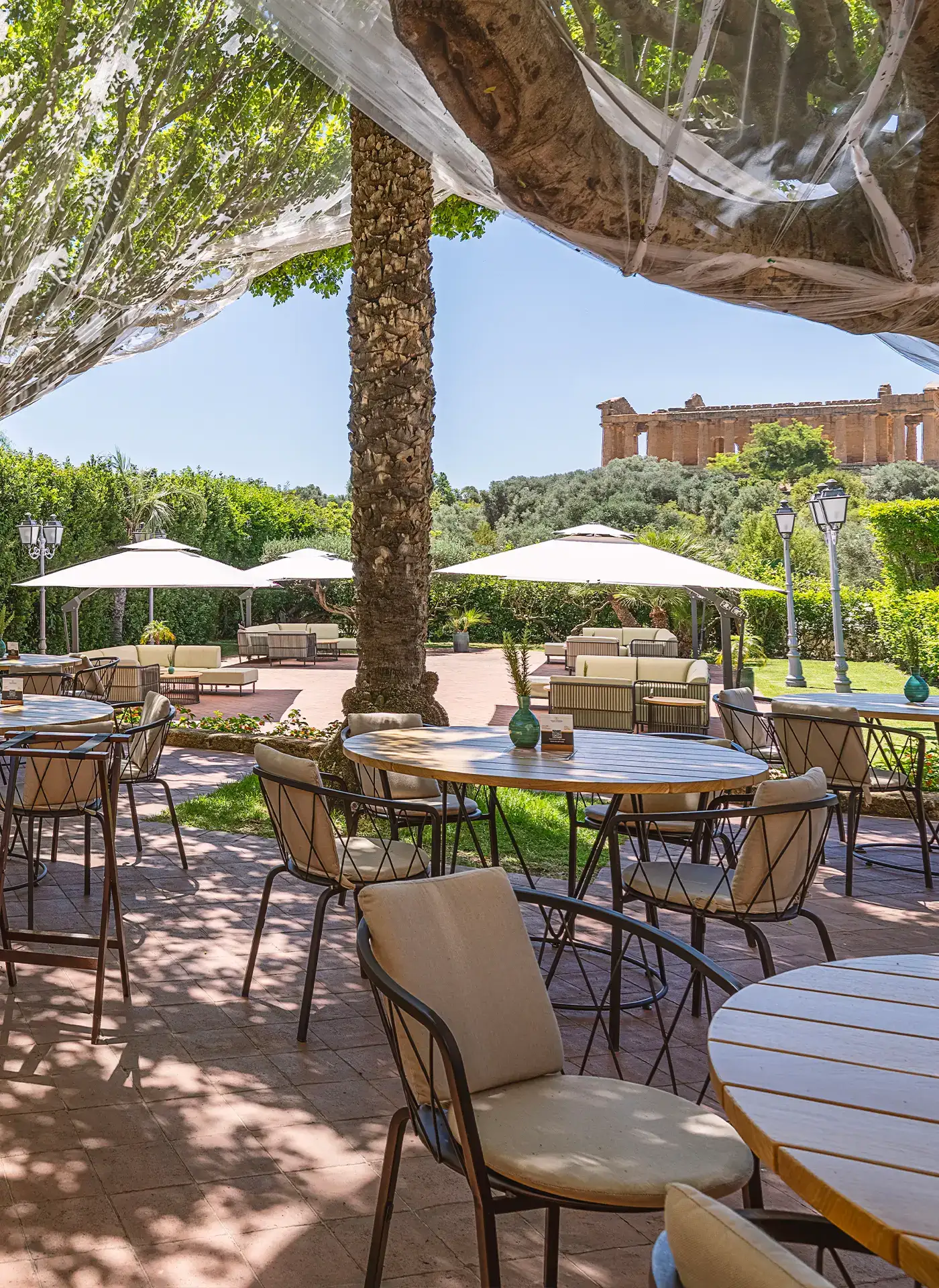 Dehor ombreggiato del Ristorante Federico II con tavoli in legno, sedie eleganti, alberi e vista panoramica sul tempio greco.