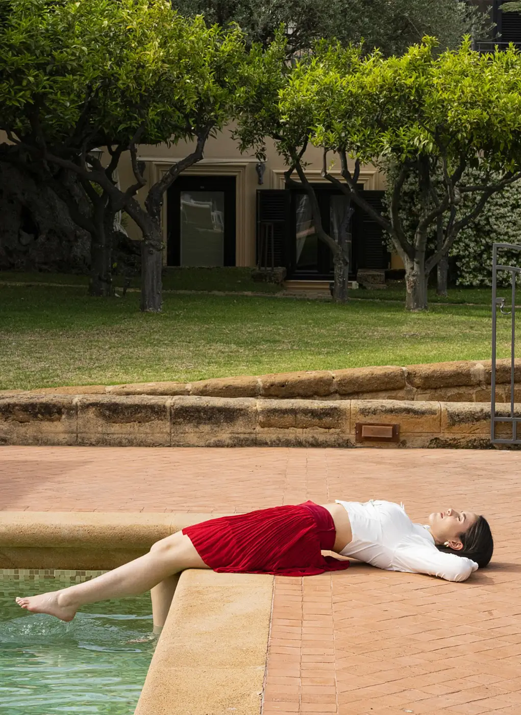 Donna sdraiata a bordo piscina presso il Villa Athena Resort, con i piedi immersi nell'acqua e giardino con alberi di agrumi sullo sfondo.