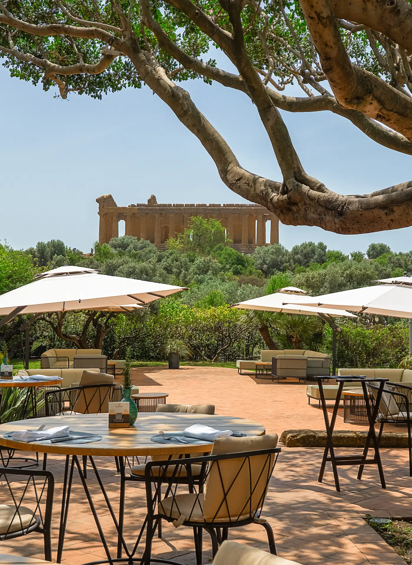 Terrazza esterna del ristorante Villa Athena con tavoli, ombrelloni e vista sul Tempio della Concordia incorniciato da rami d'albero.